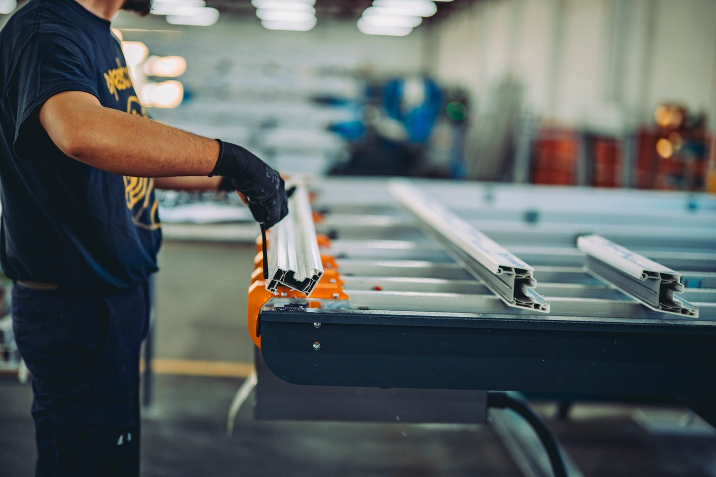 A worker in gloves setting an aluminium profile onto an assembly bench beside staged component stock.